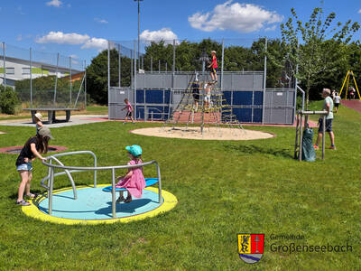 Danach durften die Kinder den neuen Spielplatz erobern. Das Karussell ist barrierefrei, daneben ist ein Bodentrampolin. Im Hintergrund vor der Multisportanlage der neue Kletterturm; rechts oben im Bild der Start der Seilrutsche. 